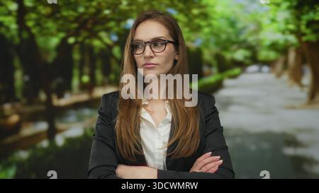Frau mit schwarzem Blazer und Brille kreuzt ihre Arme auf einem von Bäumen gesäumten Schotterweg im grünen Park; Trotz. Stockfoto