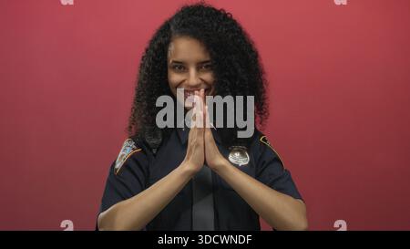Junge hispanische Polizistin in Uniform posiert lächelnd vor isoliertem roten Hintergrund und vermittelt Professionalität und Wärme Stockfoto