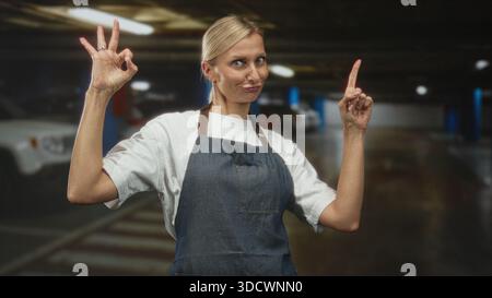Die blonde Frau im mittleren Alter mit Denim-Schürze macht mit der linken Hand ein OK-Schild und zeigt mit der rechten Hand den Zeigefinger nach oben in ein Parkhaus mit Auto Stockfoto