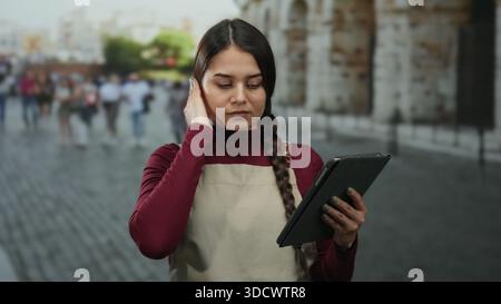 Die junge Kellnerin auf der Schürze checkt das Tablet vor dem römischen kolosseum und erscheint nachdenklich in der belebten Straßenlage mit verschwommenen Menschenmengen im Hintergrund. Stockfoto