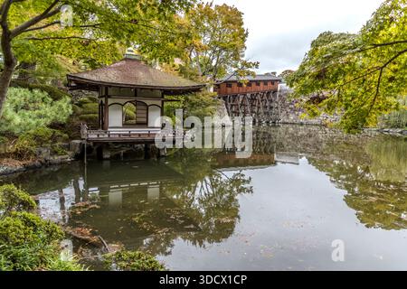 Ein traditionelles japanisches Gebäude auf Pfählen spiegelt sich im Wassergraben von Wakayama Castle, Japan, wider, während die überdachte Ohashiroka-Brücke im Hintergrund das Wasser überquert. Die Brücke bildete einst einen geschlossenen Gang, der den Nishinomaru-Garten mit den Ninomaru-Wohnräumen verband und war dem Herrn vorbehalten; ihre ungefähr elf Grad steile Neigung ist einzigartig in Japan. Der heutige Nachbau, basierend auf Zeichnungen aus der Edo-Zeit, stammt aus dem Jahr 2006. Stockfoto