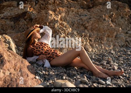 Frau, Strand, Sommer, entspannte Frau in Badeanzug mit Leopardenmuster und Strohhut, die auf Kieselstrand liegt und das Sonnenlicht der goldenen Stunde genießt Stockfoto