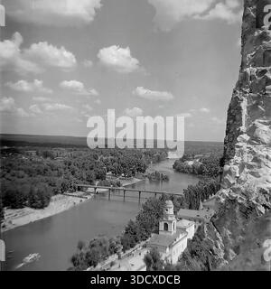 Ukraine, Region Donezk, Sviatohirsk, 1980er Jahre Ein archiviertes Schwarzweißfoto, das einen majestätischen Blick auf den Seversky Donets River von den Kreidefelsen aus aus der Luft macht. Das Bild zeigt die historischen Klostergebäude am Fuße des Berges, eine Brücke über das Wasser und ein Vergnügungsboot, das den Fluss entlang fährt. Üppige Wälder erstrecken sich bis zum Horizont unter bewölktem Himmel. Diese alte Landschaft bewahrt die malerische Schönheit des Naturschutzgebiets in einem friedlichen Donbass vor dem Krieg und diente als historisches Dokument Stockfoto