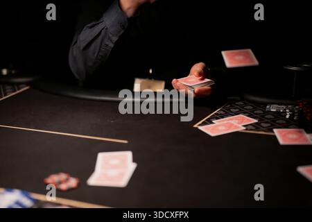 Poker-Dealer-Hand wirft Spielkarten über dunkle Casino-Tisch während Live-Kartenspiel mit Chips, Wetten-Layout und dramatischer Atmosphäre bei schlechten Lichtverhältnissen Stockfoto