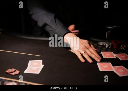 Casino Poker Tabelle Detail zeigt Dealer Hand, die Spielkarten in der Nähe von Chips und Wetten Layout, fokussierte Glücksspiel-Action im dunklen Inneren Stockfoto