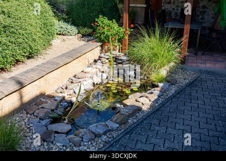 Ein ruhiger, dekorativer Gartenteich mit Wasserbrunnen, umgeben von Steinen und üppiger Vegetation, in der Nähe eines Sitzbereichs im Freien. Relaxa Stockfoto