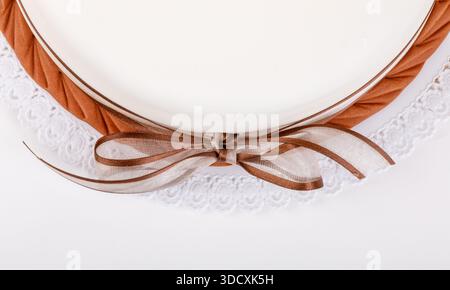 Äußerste Nahaufnahme einer leeren weißen Platte mit brauner durchscheinender Schleife, dekorativer weißer Serviettenspitze und braunem Besatz. Studio aufgenommen auf WHI Stockfoto
