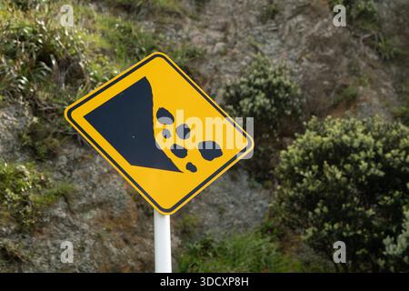 Gelb fallende Felsen Warnschild vor Felsenklippe mit Vegetation. Schwarzes grafisches Symbol für herabfallende Felsen auf gelbem Hintergrund. Rautenform. Stockfoto