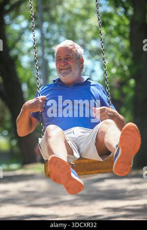 Fröhlicher Seniorenmann, der sich an sonnigen Sommertagen auf einem Spielplatz im Freien schwingt und einen verspielten Moment in einer grünen Parklandschaft genießt. Stockfoto