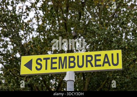 Hellgelbes Schild mit dem Text Stembureau und einem Pfeil, der einen Wahllokal für die Wahlen in den Niederlanden vor grünen Bäumen anzeigt. Stockfoto