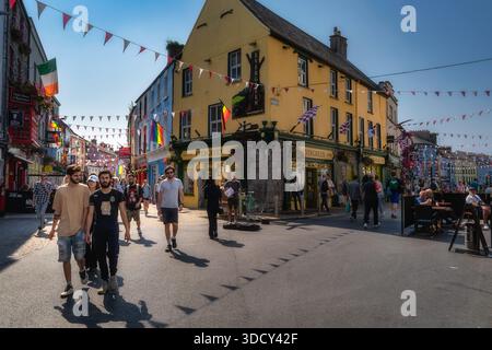 Galway, Irland, 26. August 2021 sonnendurchflutete farbenfrohe Straße mit Touristen, die den belebten Platz mit Pubs, Cafés, Festivalfladen und Sitzgelegenheiten im Freien erkunden. Paare und Gruppen, die im warmen Sommer spazieren gehen Stockfoto