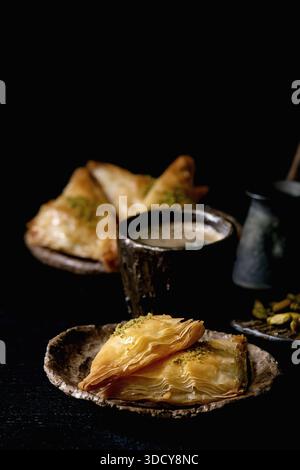 Hausgemachtes türkisches traditionelles Dessert Baklava mit Pistazien serviert auf Keramikplatte mit Nüssen, Tasse Kaffee, Vintage-Cezva auf schwarzem Holztisch. Kopierbereich Stockfoto