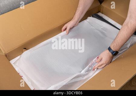 Die Hände der Person packen ein neues elektronisches Produkt aus, das noch in weißem Schaum in einer braunen Pappschachtel gesichert ist, um die frische Lieferung hervorzuheben Stockfoto