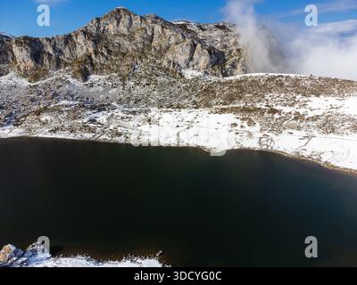 Blick aus der Vogelperspektive auf den Gletschersee Ercina mit seinen schneebedeckten Ufern und einem hohen felsigen Berg, mit Wolken, die sich erheben. Covadonga Lakes, Asturien, Spanien. Stockfoto
