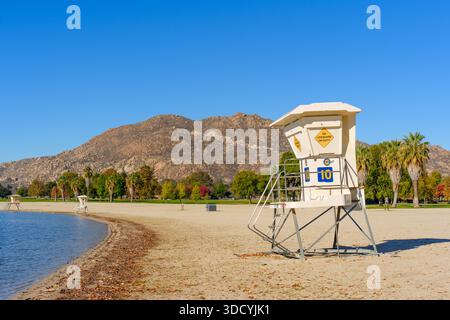 Rettungsschwimmer-Turm am Lake Perris Beach mit herbstlichen Blättern, ruhigem Seewasser, felsigen Bergen und bunten Bäumen unter einem klaren blauen Himmel. Stockfoto