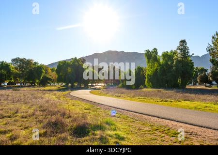 Der gepflasterte, gewundene Pfad führt durch grüne Bäume und trockenes Gras zu weit entfernten Bergen unter hellem, sonnigem Himmel in der Lake Perris State Recreation Area. Stockfoto