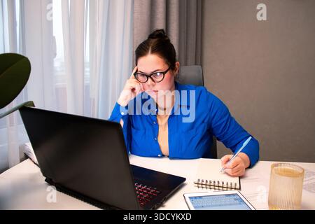 Student, der an einem virtuellen Universitätskurs per Videokonferenz teilnimmt. Junge Frau nimmt aktiv an Online-Vorträgen Teil und macht Notizen auf dem Laptop. Abstand Stockfoto