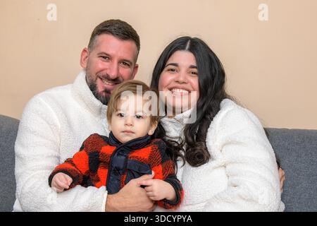 Lächelndes Familienporträt von zwei Erwachsenen, die ein Kleinkind halten, während sie drinnen zusammen sitzen. Stockfoto