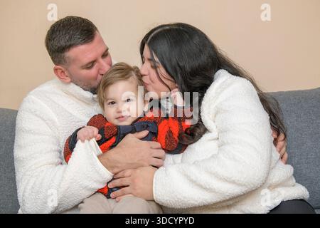 Eltern küssen ihr Kleinkind, während sie drinnen auf einer Couch sitzen. Stockfoto