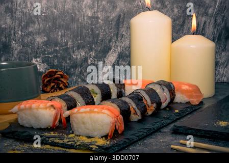 Gemischte Garnelennigiri und Sushi-Brötchen auf einem Schieferbrett mit brennenden Kerzen und Teetasse, warme japanische Abendstimmung und Entspannungskonzept. Stockfoto