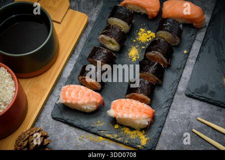 Blick von oben auf verschiedene Sushi-Brötchen und Garnelen-Nigiri auf einem Schieferbrett mit Sojasauce und Reisschalen, elegante japanische Tischdekoration. Stockfoto