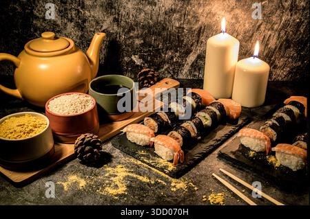 Japanisches Dinner Stillleben mit Sushi-Brötchen, Nigiri, Teekanne, Reisschalen und Kerzen auf einem dunklen Steintisch, gemütliches Gourmet-Restaurant Ambiente. Stockfoto
