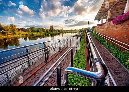 Eine zickzackige Steinrampe für Behinderte auf einem Stadtboulevard am Fluss. Praktische Stahlhandläufe. Ein heller Sommertag mit einem wunderschönen Himmel. Stockfoto