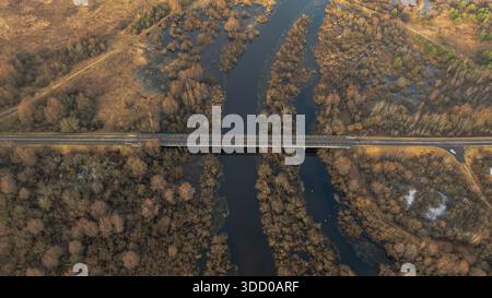 Eine zweispurige Brücke in Belarus führt über einen sich windenden Fluss durch Sumpfgebiete und baumlose Wälder im Winter, mit einem Fahrzeug direkt drin Stockfoto