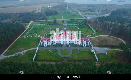 Aus der Vogelperspektive eines neogotischen Palastes in Belarus mit weißen Wänden und roten Dächern, gelegen in einem symmetrischen ummauerten Garten, dichtem Wald, offenen Feldern, See und Stockfoto