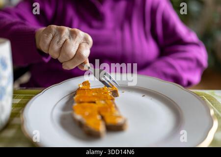 Wlderly Woman sitzt an einem Tisch und bereitet ein Stück Brot mit Gelee mit einer Gabel zu essen. Sie trägt einen lila Pullover und genießt zu Hause Essen Stockfoto