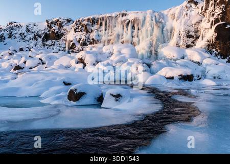 Winterszene im Freien mit einem schneebedeckten Fluss in der Nähe des teilweise gefrorenen Oxararfoss Wasserfalls im Thingvellir Nationalpark im Goldenen Kreis in Island. Stockfoto