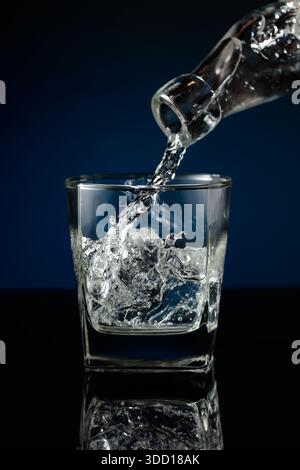 Kristallklarer Wodka oder Gin wird über Eis in ein Glas gegossen. Wodka mit Eis auf schwarzem reflektierendem Hintergrund. Stockfoto
