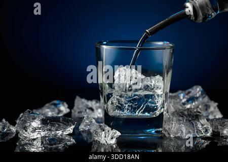 Kristallklarer Wodka oder Gin wird über Eis in ein Glas gegossen. Wodka und Eisstücke auf schwarzem reflektierendem Hintergrund. Kopierbereich. Stockfoto