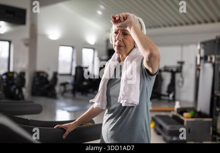 Eine ältere müde Frau wischt sich den Schweiß von der Stirn, während sie im Fitnessstudio ein Laufband benutzt. Sie konzentriert sich auf ihre Fitness-Routine im Morgenlicht. Stockfoto