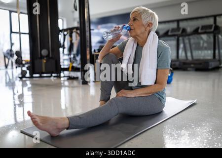 Eine Frau sitzt auf einer Matte im Fitnessstudio und trinkt nach dem Training Wasser. Sie hat ein Handtuch um den Hals und sieht entspannt aus. Stockfoto