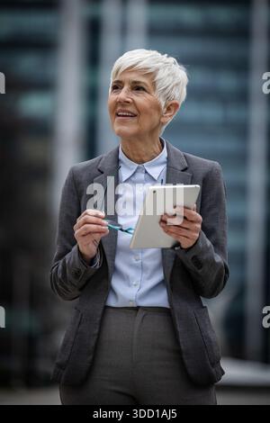 Eine Frau mit kurzen grauen Haaren steht draußen in Geschäftskleidung. Sie hält ein Tablet in der Hand und scheint etwas zu reflektieren oder zu planen. Stockfoto