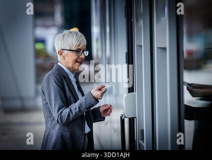 Eine Frau im Anzug hält eine Karte, während sie versucht, ein Gebäude oder einen geldautomaten zu betreten. Sie sieht glücklich aus und steht vor der Tür Stockfoto