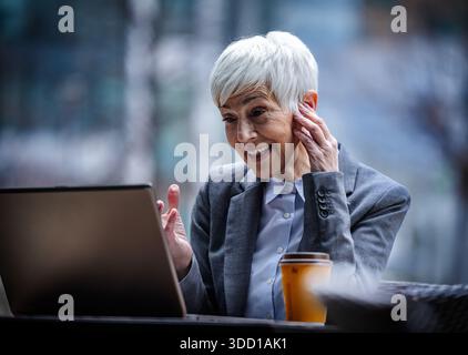 Geschäftsfrau sitzt an einem Tisch im Café im Freien und hat Videoanrufe auf dem Laptop. Sie trägt einen Anzug und hat kurze Haare. Stockfoto