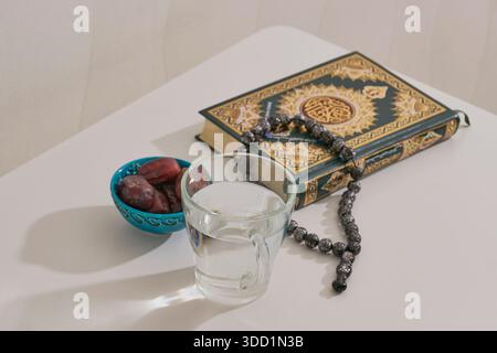Traditionelle Iftar-Einrichtung, um nach Sonnenuntergang während des Ramadan schnell zu brechen. Glas Wasser und Datteln in türkisfarbener Schüssel neben dem Heiligen Koran und einer Gebetsreihe Stockfoto