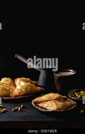 Hausgemachtes türkisches traditionelles Dessert Baklava mit Pistazien serviert auf Keramikplatte mit Nüssen, Tasse Kaffee, Vintage-Cezva auf schwarzem Holztisch. Kopierbereich Stockfoto