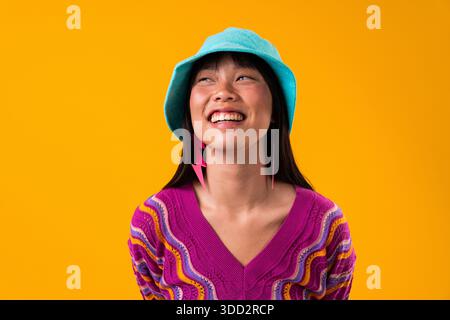 Wunderschönes Porträt einer chinesischen Frau auf farbenfrohem Hintergrund. Lächelnde junge asiatische Frau posiert in einem Atelier, trägt einen lebendigen Eimerhut und einen lila Pullover Stockfoto