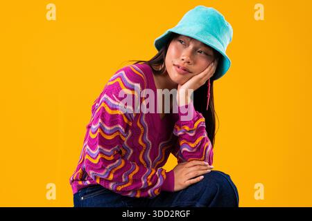 Wunderschönes Porträt einer chinesischen Frau auf farbenfrohem Hintergrund. Eine junge asiatische Frau mit blauem Eimerhut und einem pulverförmigen Pullover, die nachdenklich-fu posiert Stockfoto