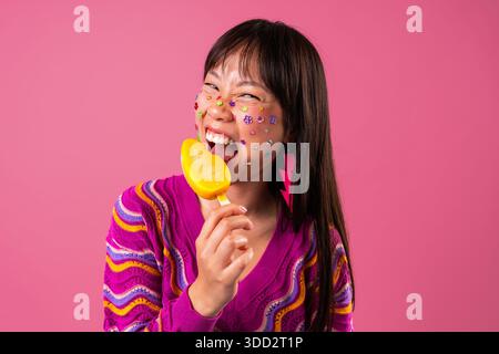 Wunderschönes Porträt einer chinesischen Frau auf farbenfrohem Hintergrund. Junge asiatische Frau, die fröhlich ein gelbes Eis vor rosa Hintergrund isst, Gesichtsjuwelen, Farbe Stockfoto