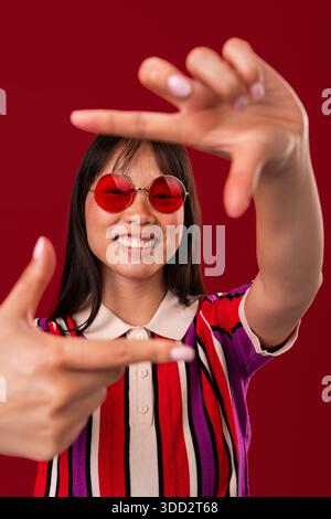 Wunderschönes Porträt einer chinesischen Frau auf farbenfrohem Hintergrund. Lächelnde asiatische Frau, die ihr Gesicht mit Händen umrahmt, ein buntes gestreiftes Hemd und eine rote Runde trägt Stockfoto