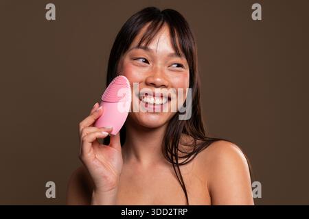 Wunderschönes Porträt einer chinesischen Frau auf farbenfrohem Hintergrund. Junge asiatische Frau lächelt, während sie eine rosa Silikon-Gesichtsreinigungsbürste für die Hautpflege verwendet Stockfoto