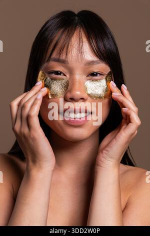 Wunderschönes Porträt einer chinesischen Frau auf farbenfrohem Hintergrund. Junge asiatische Frau lächelt, trägt luxuriöse goldene Augenflecken für Schönheit auf, Anti-Aging Stockfoto