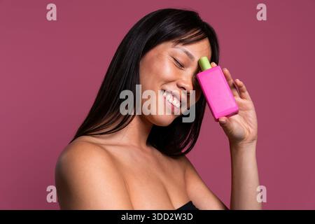 Wunderschönes Porträt einer chinesischen Frau auf farbenfrohem Hintergrund. Asiatische Frau lächelt mit geschlossenen Augen und hält eine pinkfarbene Flasche Schönheitsprodukt gegen sie Stockfoto