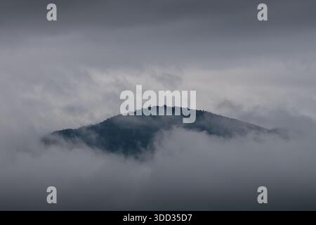 Ein einsamer, nebelumhüllter Berggipfel, der aus einem Meer dunkler, brütender Wolken auftaucht und ein Gefühl von Geheimnis, Isolation und atmosphärischem Drama vermittelt. Stockfoto