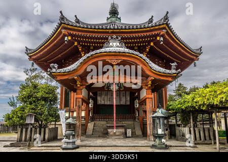 Die kunstvoll geschnitzten Dachvorsprünge der achteckigen, rotlackierten Halle des Kofuku-ji-Tempels erheben sich vor einem bewölkten Himmel in Nara, Japan, flankiert von Stehlaternen am Fuße der Stufen – wahrscheinlich der Nan'endo (Südliche Achteckhalle), der neunte Halt auf der Saigoku-Kannon-Pilgerroute. Die aktuelle, vierte Halle stammt aus dem Jahr 1789 und beherbergt die Fukukensaku-Kannon-Statue, einen Nationalschatz. Stockfoto
