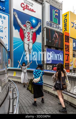 Eine Frau posiert mit ausgestreckten Armen vor der Glico-Werbetafel am Dotonbori-Kanal in Osaka, Japan, während zwei Begleiter sie fotografieren. Sie ahmt die Pose des laufenden "Glico Man" nach. Der Süßwarenhersteller Ezaki Glico hat die erste Version des Schilds 1935 aufgehängt. Ursprünglich warb es für einen Energieriegel mit Glykogen aus Austern, der Ursprung des Markennamens "Glico". Die aktuelle LED-Version ist seit 2014 beleuchtet. Stockfoto
